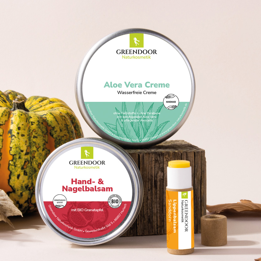 Drei Greendoor Produkte vor herbstlichem Hintergrund mit einem Kürbis und Holz. Zu sehen sind Greendoor Handbalsam, Aloe Vera Creme und Lippenbalsam